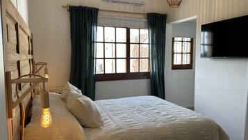 Une chambre à coucher comprenant un lit, une lampe de chevet, une fenêtre avec des rideaux, un appareil de climatisation et un téléviseur fixé au mur.