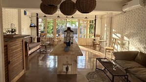 Un salón amplio con suelo de madera, un sofá, una mesa de centro y una mesa de comedor. Hay lámparas de mimbre colgando del techo y una pared de ladrillo al fondo.