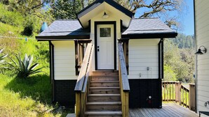 A wooden deck leading to a small house with a door and a window, surrounded by greenery.