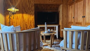 Una habitación acogedora con paredes de madera, un televisor, una mesita con lámpara y dos sillas de madera.