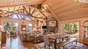 A spacious log cabin with a high ceiling, a fireplace, a dining area, and large windows offering views of the surrounding forest. - Amazing Lakefront Getaway-sleeps 18 guests, w/private dock, gameroom, spa & more (Hayden)