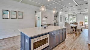 A modern kitchen with a central island, pendant lights, and a view into the dining area. - Luxury Beach Oasis~Sleeps 26! FREE Golf Cart~Pool/HotTub~Game Room (Destin)