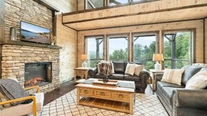 Un salon chaleureux avec une cheminée en pierre, des meubles en bois et de grandes fenêtres donnant sur l’extérieur.
