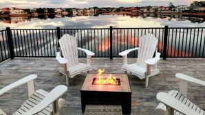 Un patio con sillas Adirondack y un pozo de fuego, con vista a una masa de agua tranquila y una costa lejana.