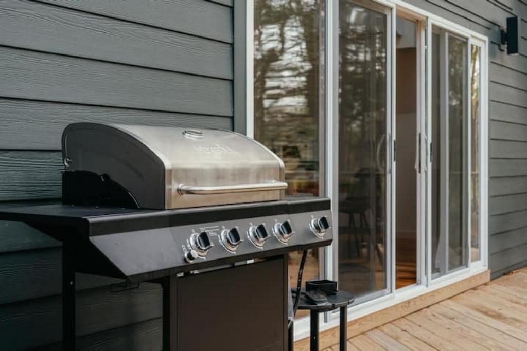 Una barbacoa d'acer inoxidable amb quatre fogons, situada sobre un terra de fusta davant d'una casa de revestiment gris i amb una porta corredissa de vidre.