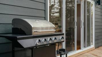 Una barbacoa d'acer inoxidable amb quatre fogons, situada sobre un terra de fusta davant d'una casa de revestiment gris i amb una porta corredissa de vidre.
