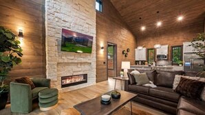 A modern living room with a stone fireplace, a sofa, a coffee table, and a kitchen area visible through an open door.