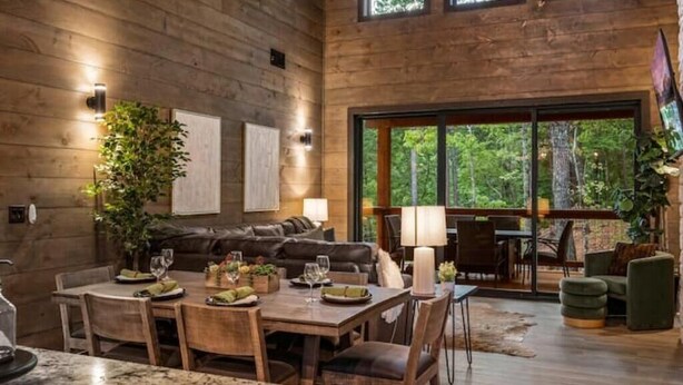 A spacious living area with a wooden floor, a dining table set with plates and glasses, a large window offering a view of the outdoors, and a cozy seating area with a sofa and chairs.