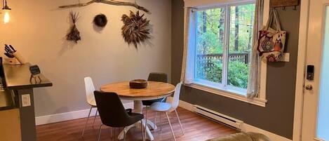 A dining area with a round wooden table, two black chairs, and a white chair. There is a window with a view of greenery outside.