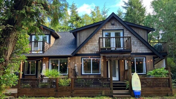 A two-story wooden house with a balcony, a porch with string lights, and a small staircase leading to the entrance. - Sienna's Tree House #3 (Tofino)