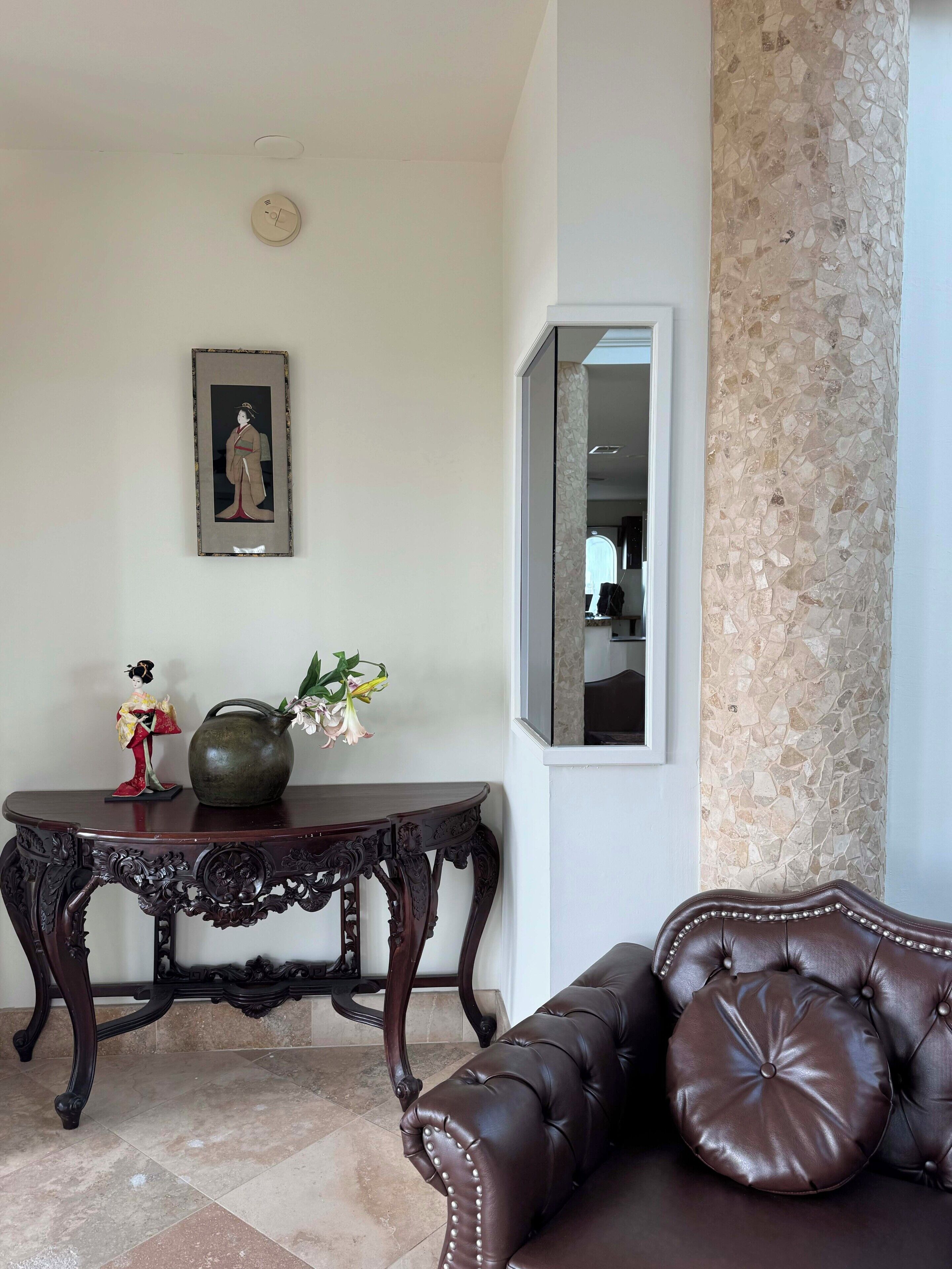 A living room with a dark brown leather sofa, a wooden side table with a vase and figurine, a framed artwork on the wall, and a textured column.