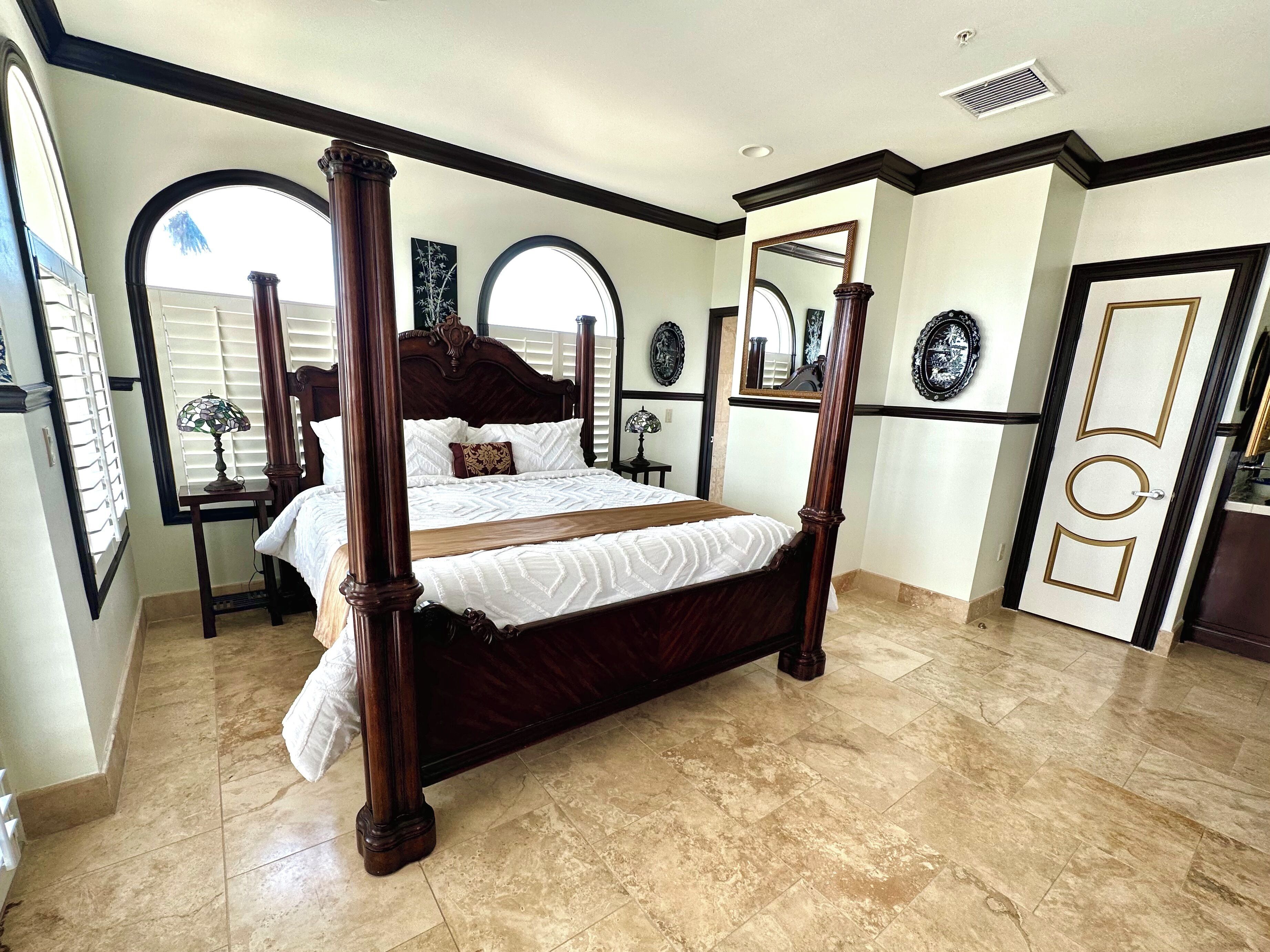 A four-poster bed with a dark wooden frame and white bedding, a large arched window, and a door with decorative panels.