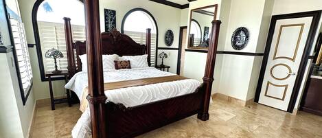 A four-poster bed with a dark wooden frame and white bedding, a large arched window, and a door with decorative panels.