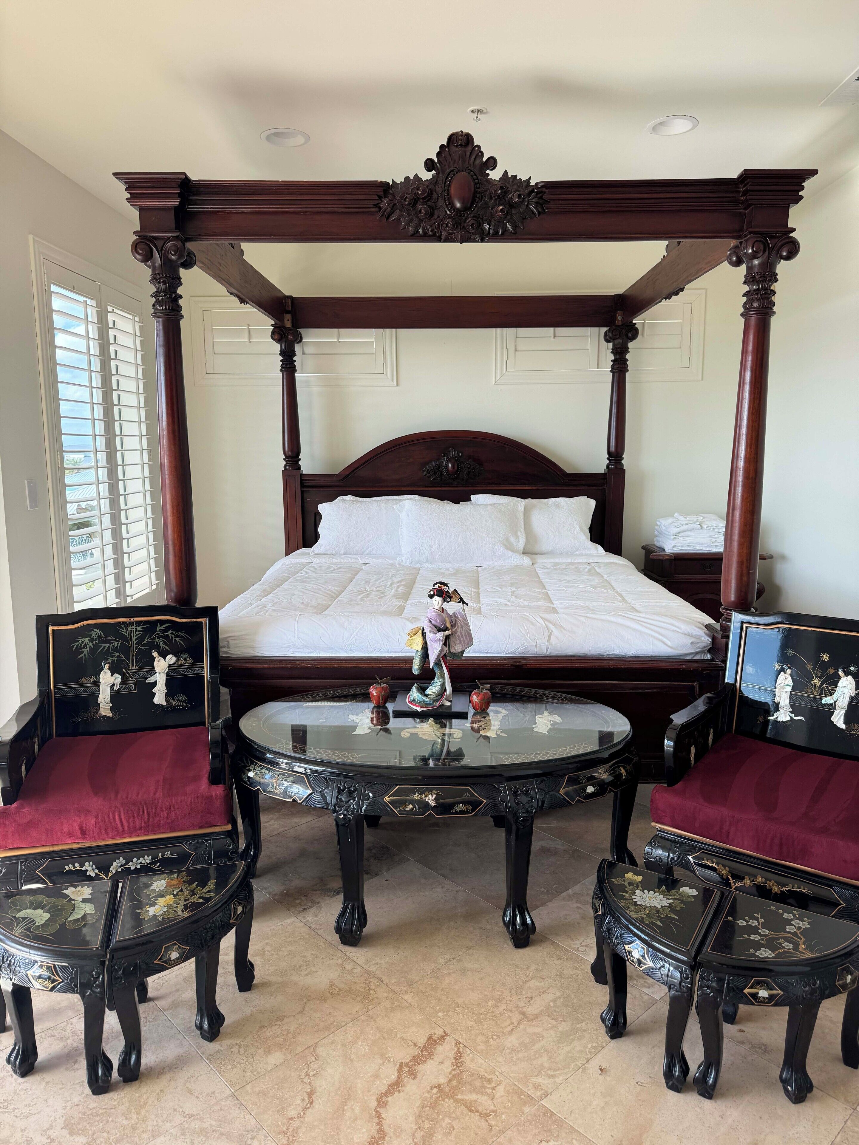 A four-poster bed with a decorative headboard, two round tables with chairs, and a window with blinds.