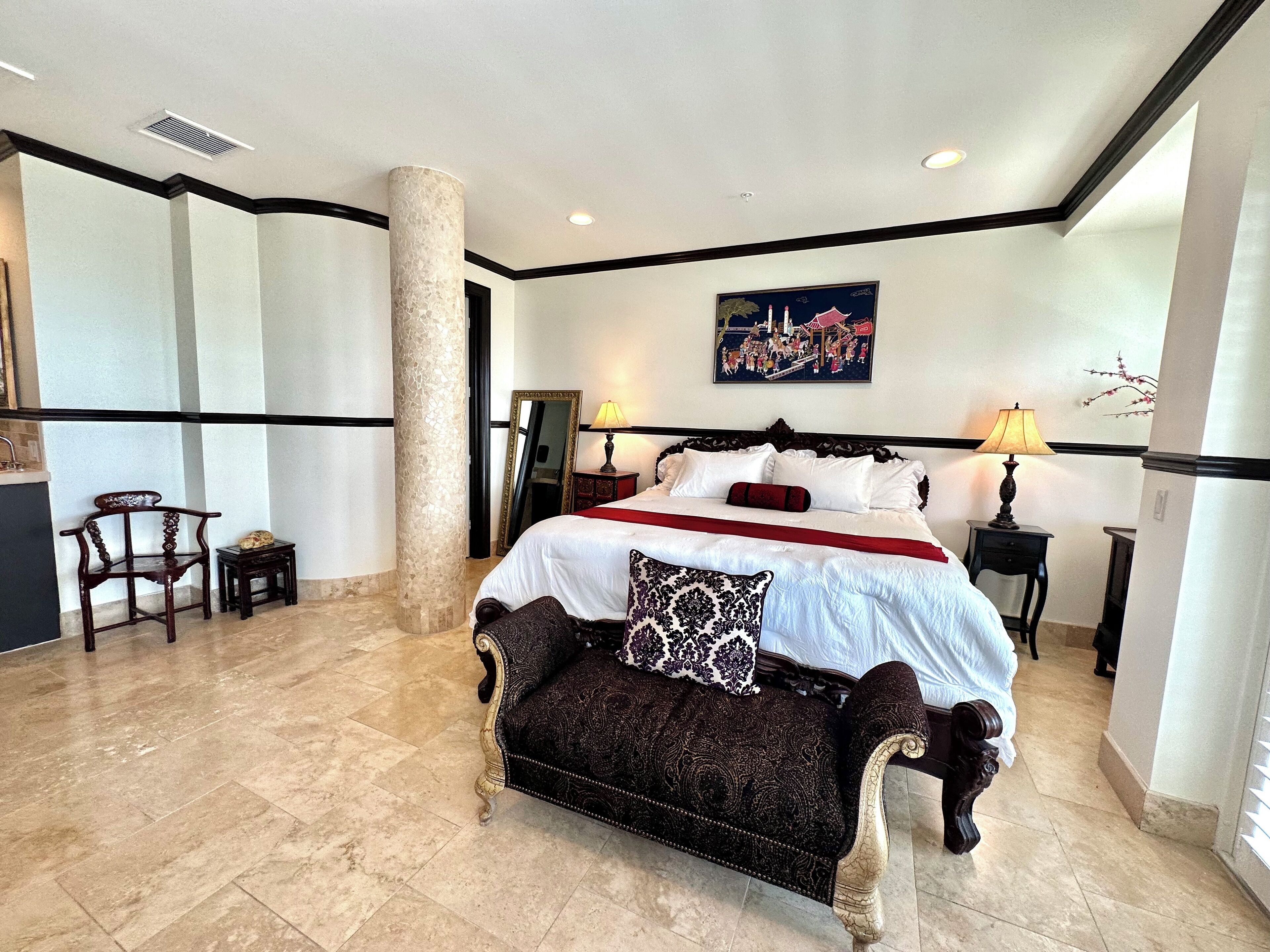 A hotel room with a large bed, two bedside tables with lamps, a chair, a large mirror, and a painting on the wall.