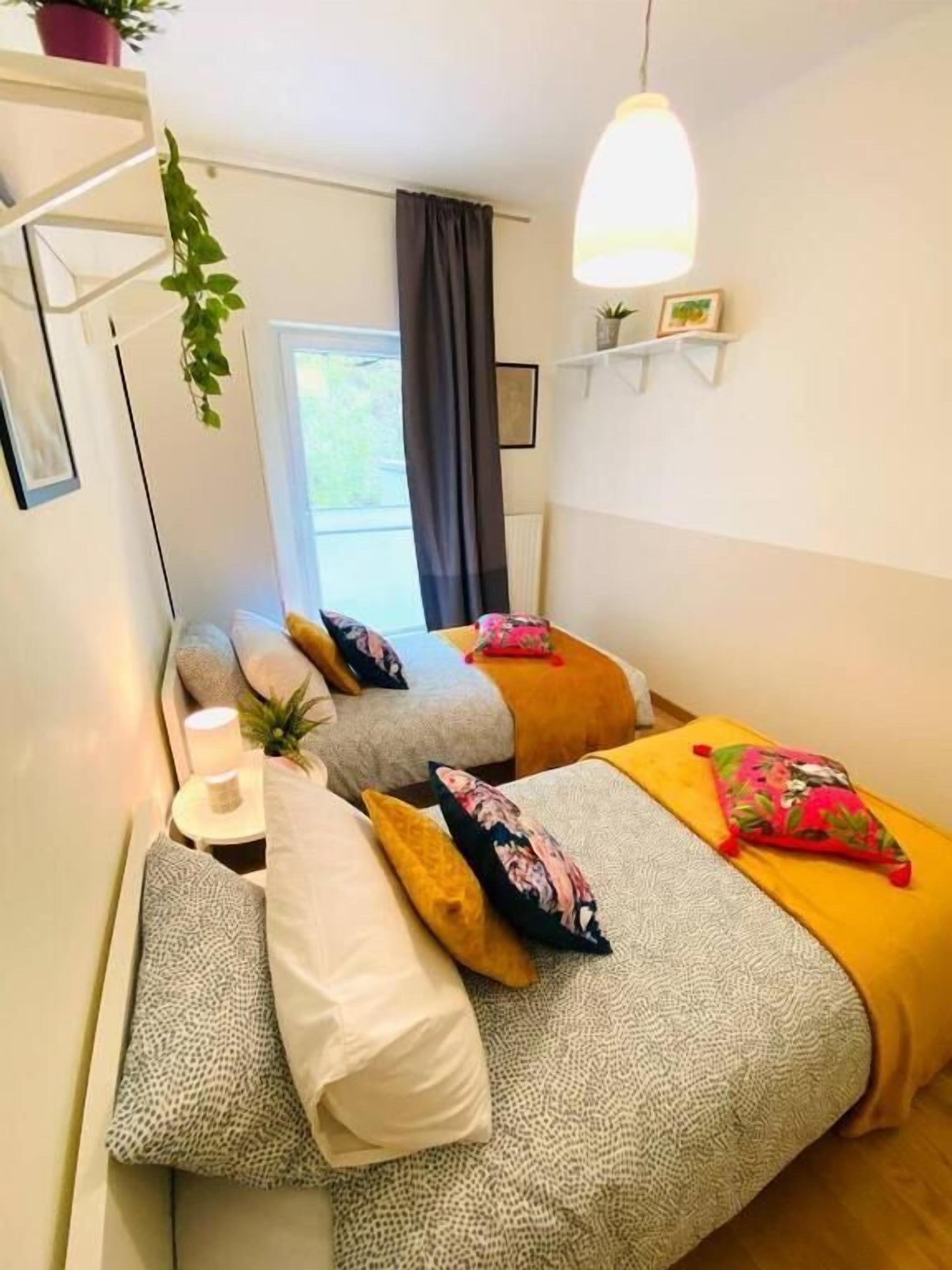 Een slaapkamer met twee bedden, een nachtkastje met een lamp, een hangplant en een raam met gordijnen.