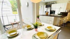 A modern dining area with a long table set for four, a hanging light fixture, and a view of the kitchen in the background. - Large 3bedrooms with Terrace and Parking (Luxembourg City)