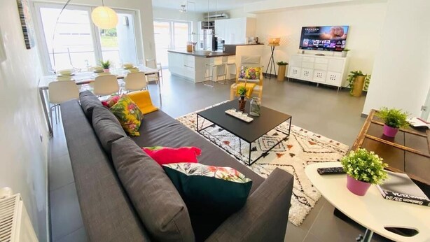 A modern living room with a sofa, coffee table, and TV. There is a kitchen in the background with a TV and cabinets. - Large 3bedrooms with Terrace and Parking (Luxembourg City)