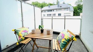 A balcony with a wooden table, two chairs, a potted plant, and a view of buildings in the background. - Large 3bedrooms with Terrace and Parking (Luxembourg City)
