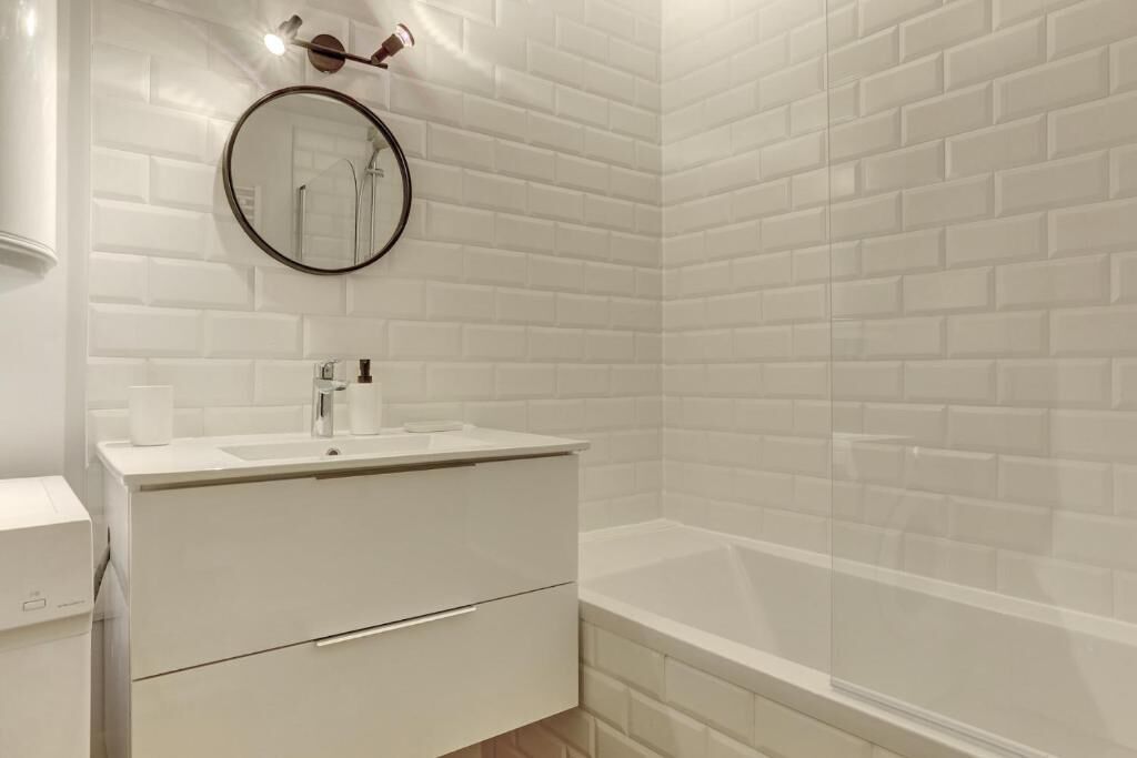 Un baño moderno con azulejos blancos tipo metro, un espejo redondo, un lavabo blanco y una bañera.