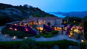 Ein großes, zweistöckiges Haus mit Naturstein-Fassade, umgeben von gepflegten Gärten und Blick auf das Meer.