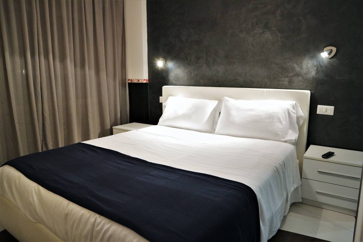 A neatly made bed with white linens and a dark blue accent, a white nightstand with a remote control, and a dark textured wall.