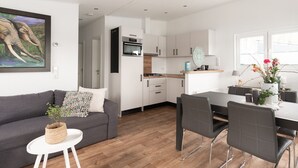 Een moderne keuken met witte kasten, een eethoek met een tafel en stoelen, een grijze bank en een klein wit bijzettafeltje met een plant.