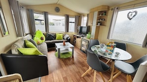 Un salon confortable avec un canapé, une table basse et une table à manger avec des chaises. On y trouve également un téléviseur et une étagère remplie de divers objets.