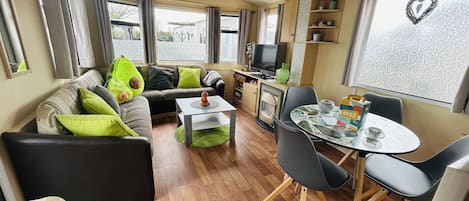 A cozy living area with a sofa, coffee table, and a dining table with chairs. There is a television and a shelf with various items.