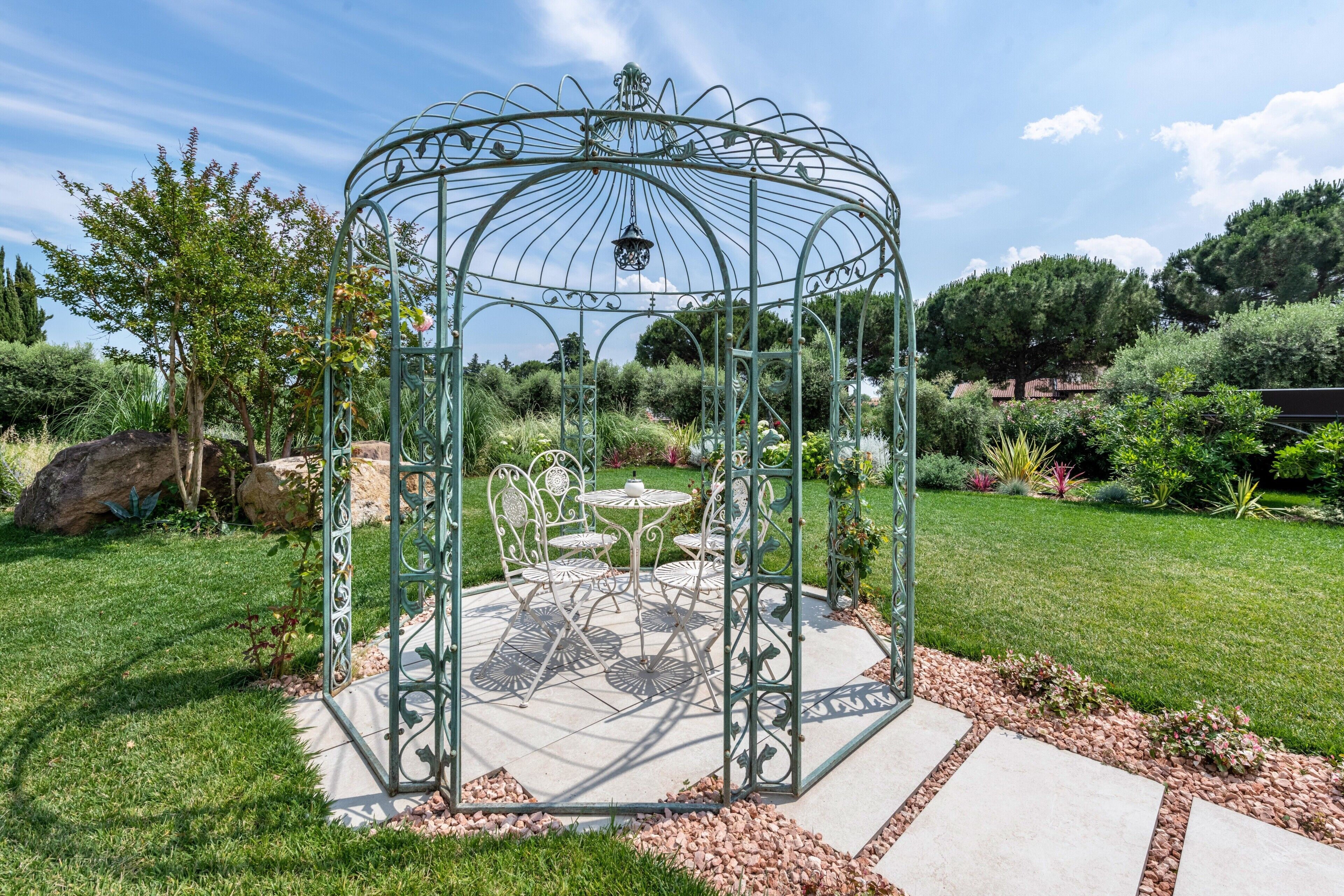Gazebo metàl·lic amb taula i cadires, envoltat d'un jardí ple de plantes variades i un cel clar.
