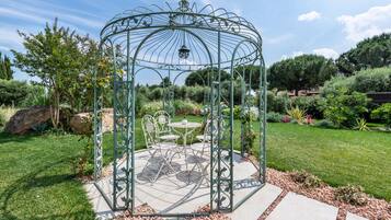Gazebo metàl·lic amb taula i cadires, envoltat d'un jardí ple de plantes variades i un cel clar.