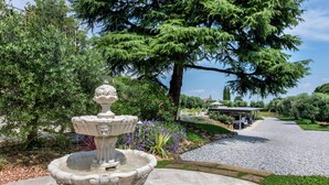 Ein Steinkunstbrunnen mit dekorativer Oberseite, umgeben von üppigem Grün und einem Kiesweg, der zu einem überdachten Gebäude führt.