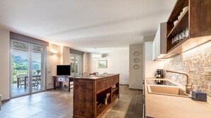 A modern kitchen with a large island, a built-in oven, and a sink. There is a dining area with a table and chairs, and a sliding glass door leading to a balcony. - Apartment 'Abaye 6 Cabernet - Charm' with Shared Pool, Wi-Fi and Air Conditioning (Soiano del Lago)