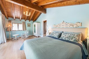 Une chambre à coucher avec un grand lit, des poutres apparentes en bois au plafond, un lustre, une commode, une chaise et une fenêtre avec des rideaux.