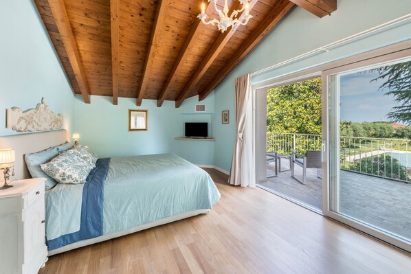 Une chambre avec un plafond en bois, un lit recouvert d’une courtepointe bleue, un téléviseur fixé au mur et un balcon aménagé avec du mobilier d’extérieur.
