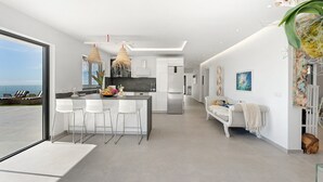 Een moderne keuken met een centraal kookeiland, barstoelen, een hangende lamp en uitzicht op de oceaan.