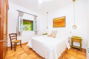 Ein Schlafzimmer mit einem Bett, einem Sessel, einem Fenster mit Vorhängen, einem Nachttisch und einem Bild an der Wand.