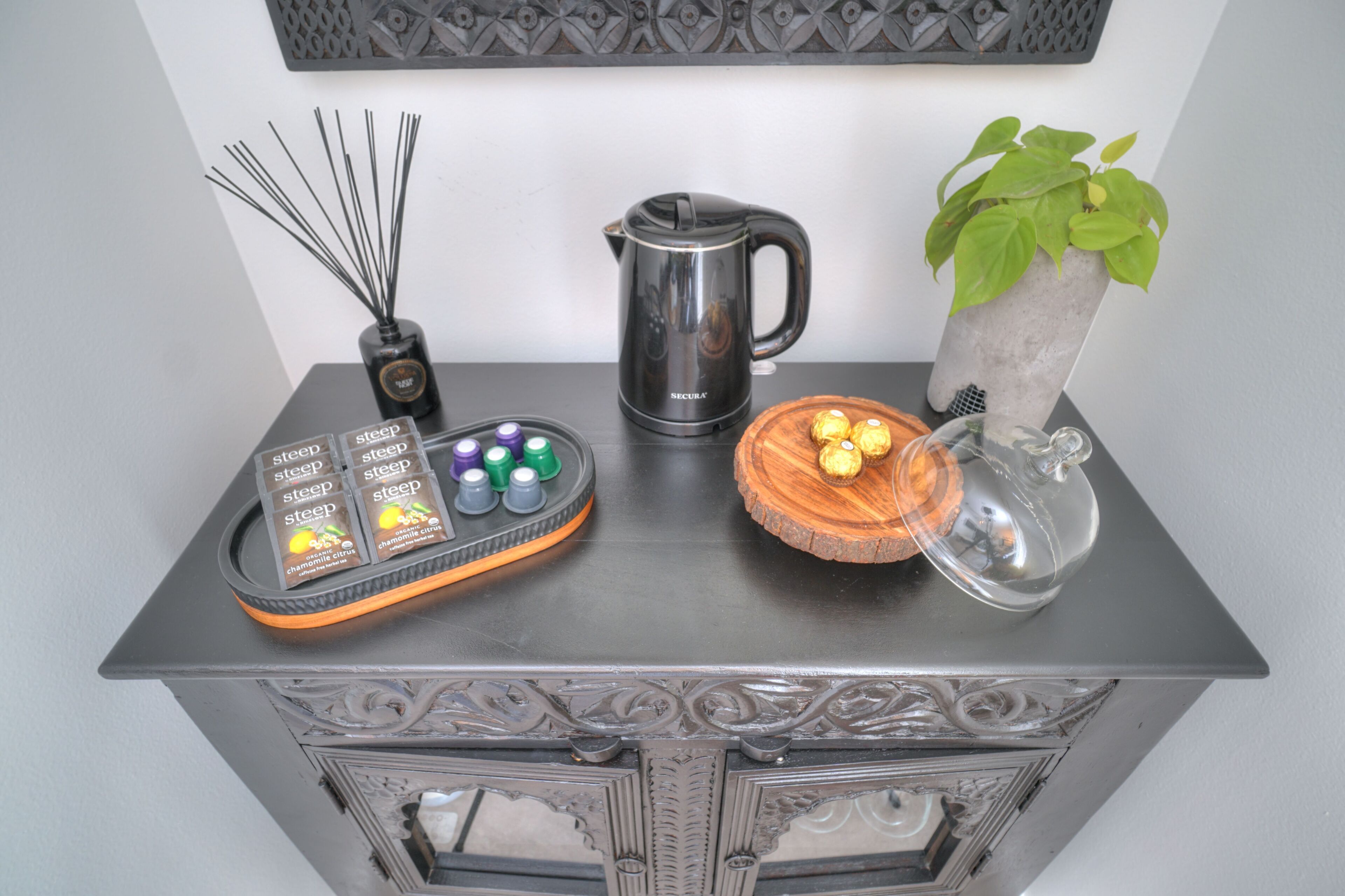 A kitchen counter with a coffee maker, a diffuser, a tray with chocolates, and a potted plant.