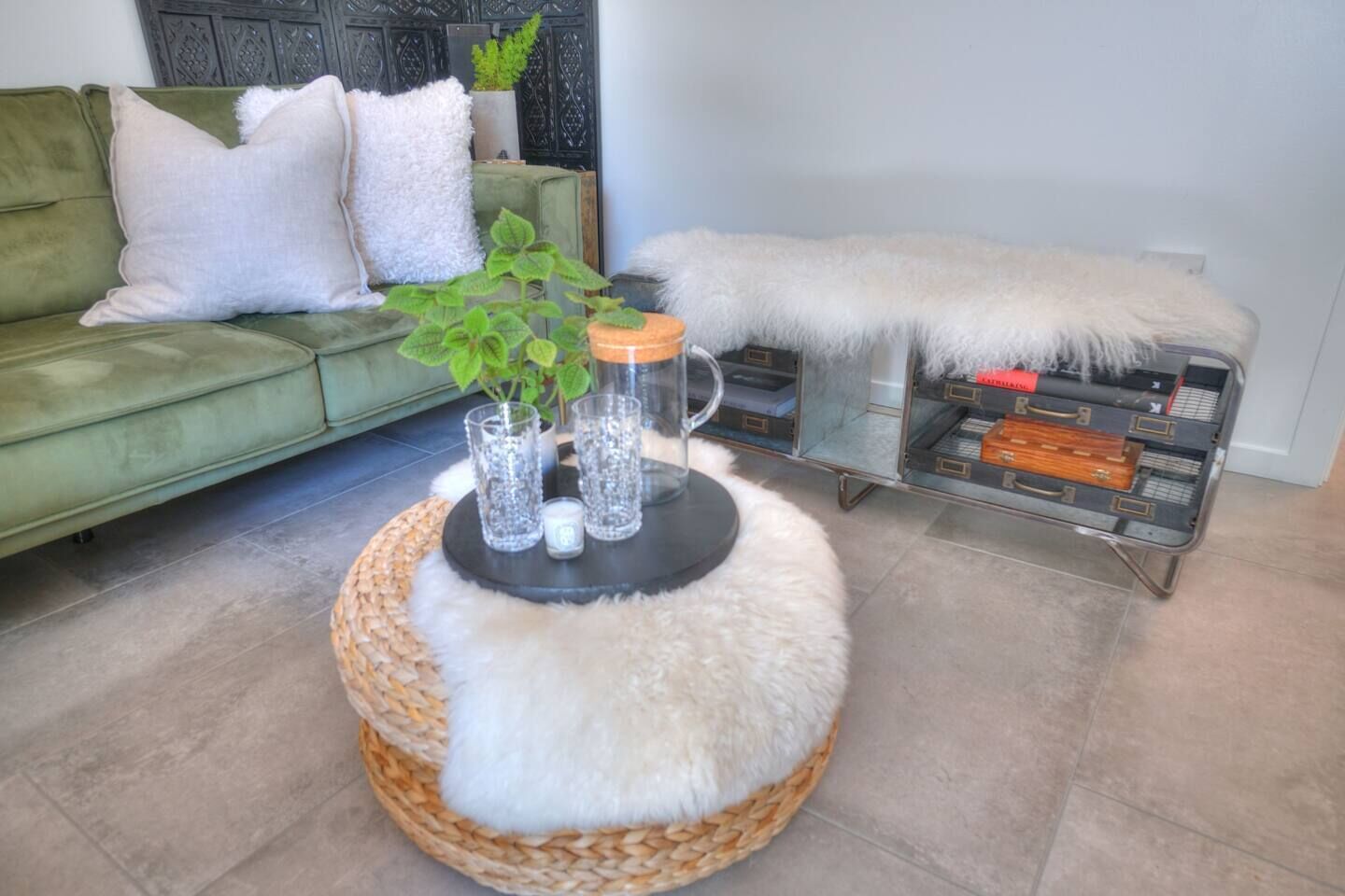 A living room with a green sofa, a round coffee table with a white furry ottoman, a glass side table with decorative items, and a wicker basket.