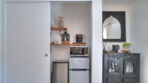 Uma cozinha compacta com micro-ondas, geladeira e uma pequena lixeira. Há um armário decorativo com um espelho acima.