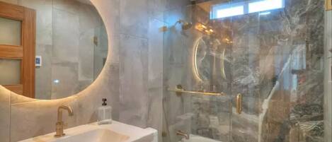 A bathroom with a large oval mirror, a white sink, a glass-enclosed shower, and marble walls.