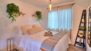 Quarto com cama, mesa de cabeceira, planta pendente, janela com cortinas e escada com plantas em vasos.