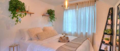 A bedroom with a bed, a bedside table, a hanging plant, a window with curtains, and a ladder with potted plants.