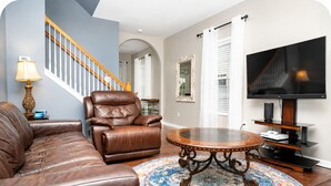 Ein Wohnzimmer mit einem braunen Ledersofa, einem Ledersessel, einem runden Couchtisch und einem Teppich mit Muster. Ein Fernseher und eine Treppe mit Holzwandleiste sind ebenfalls zu sehen.