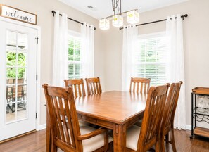 Une salle à manger avec une table en bois et six chaises, un lustre et une pancarte « Bienvenue » au-dessus d’une porte.