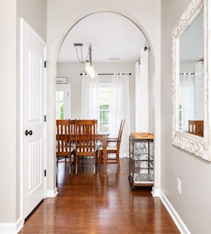 A hallway with a wooden floor, white walls, a white door, a dining area with wooden chairs and a table, and a large mirror on the wall.