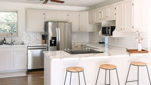 A modern kitchen with white cabinets, stainless steel appliances, a marble island with bar stools, and a ceiling fan. - The Kennewick Cottage  (Kennewick)