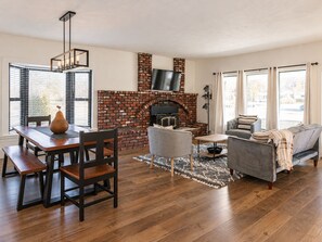 A living room with a brick fireplace, a dining table with chairs, a sofa, and a television mounted above the fireplace. - The Storybook Cottage (Kennewick)