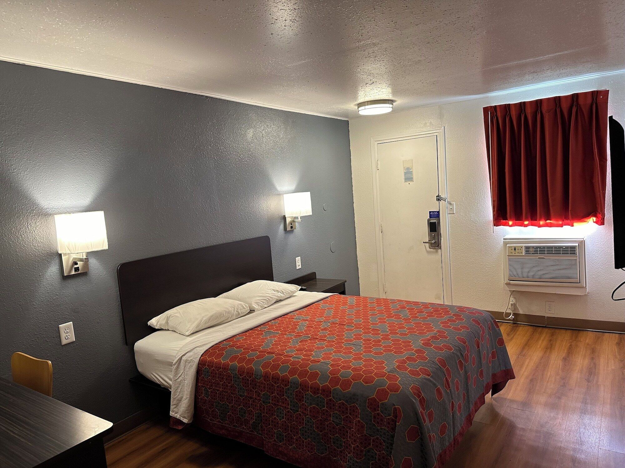 A hotel room with a bed, two bedside lamps, a door with a lock, a window with red curtains, and an air conditioning unit.