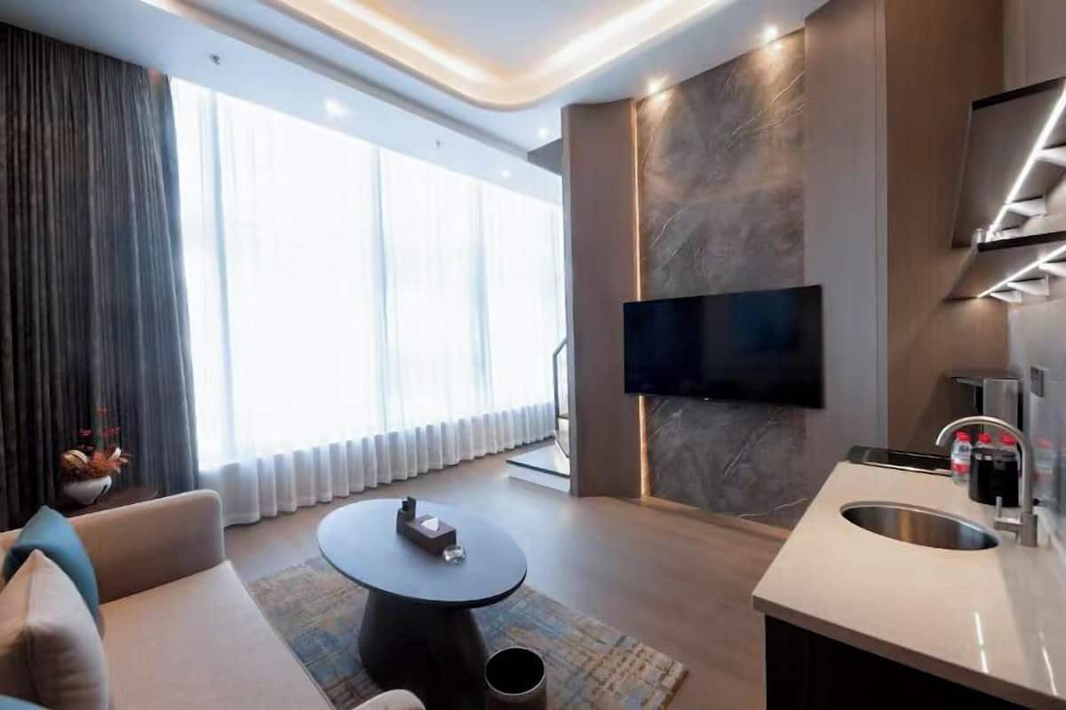 Ett modernt hotellrum med en stor, platt-TV som är monterad på väggen, en soffa, ett runt soffbord, en liten köksdel med ett handfat och ett stort fönster med gardiner.